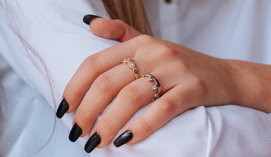 A woman's hand adorned with two elegant rings featuring intricate designs, paired with black manicured nails.